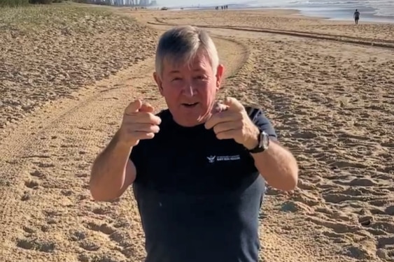 70-year-old man kneeling on beach in blue shorts and shirt and pointing at camera.