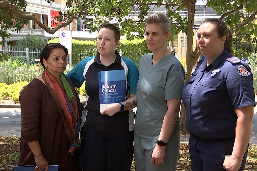 a group of four women, four of whom are health professionals, standing outdoors and speaking to the press