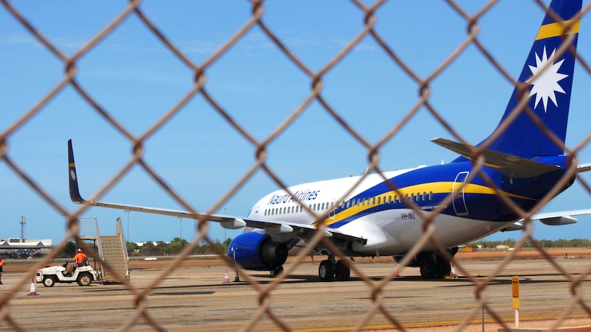A Nauru Airlines plane sits on an airpot tarmac, viewed through cyclone fencing.