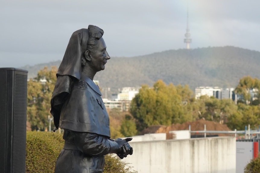 The statue is of a woman in nursing clothing, her hands clasped, Telstra Tower and a rainbow in the background.