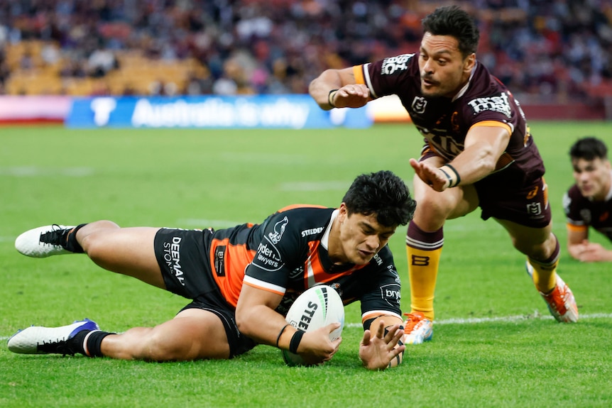 A Wests Tigers NRL player grounds the ball as he slides in to score a try against the Broncos.