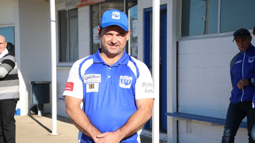 A man in a blue cap and football shirt stands in front of football club rooms.