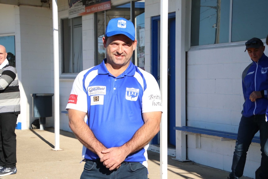 A man in a blue cap and football shirt stands in front of football club rooms.