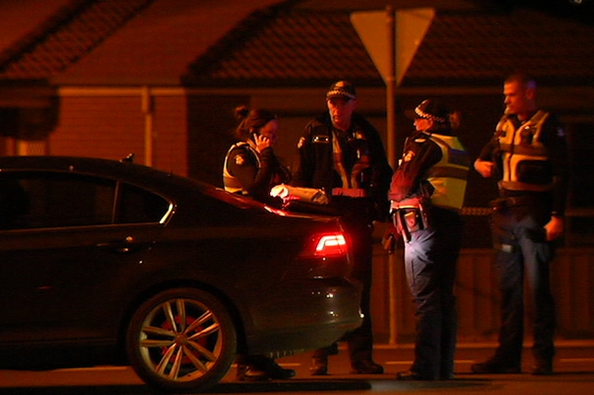A group of four Victoria Police officers standing next to a dark coloured car on a street at night time.