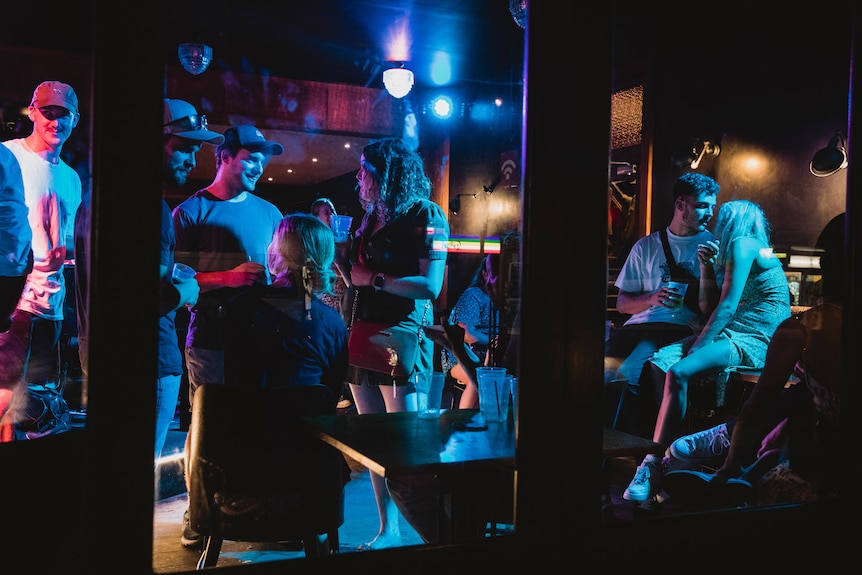 A group of young people hang out inside a bar with blue light overhead.