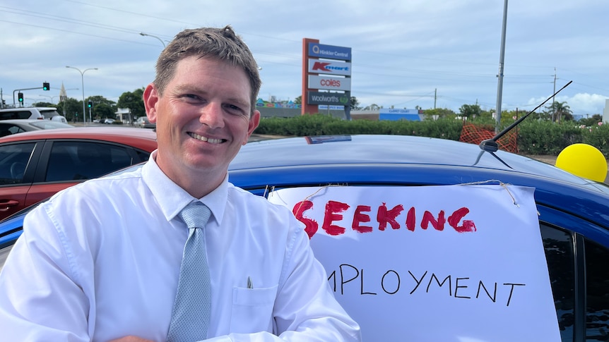 A young man stands in front of a blue car with his arms crossed next to a sign that reads 'seeking employment now'. 