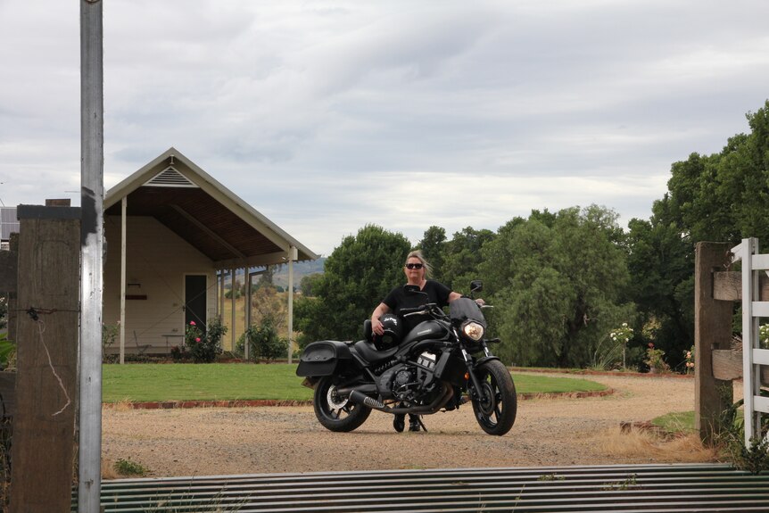 A middle-aged blonde woman in all black clothing standing behind a black motorcycle in front of a country home.