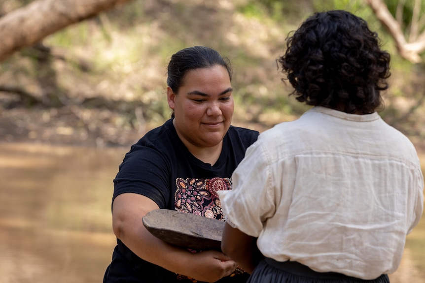 Woman hands a hat to another woman near a lake 