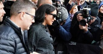 Meghan Markle wears a black coat and sunglasses and holds a grey bag as she is surrounded by paparazzi.