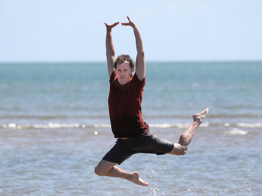 A man jumps out of the water at the beach in a dancer's pose.