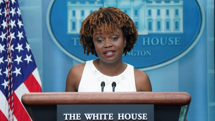 A black woman with a bob-style haircut stands at a lecturn labelled as The White House, in front of an American flag