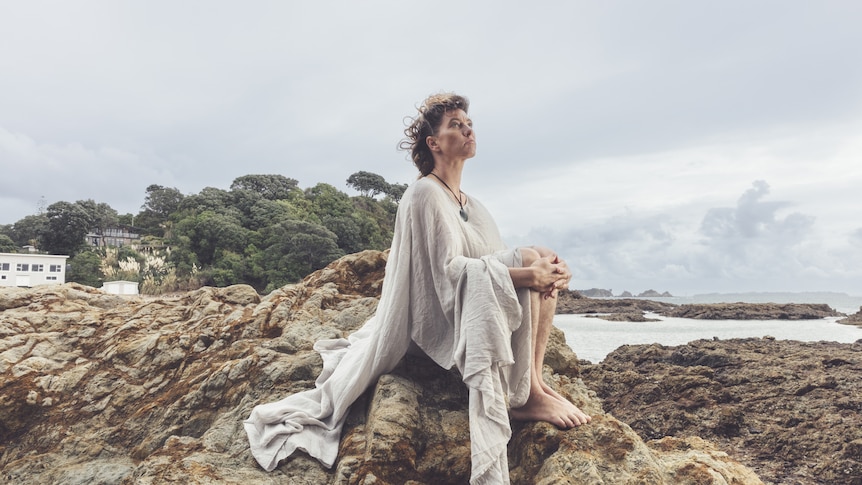 Amanda Palmer sits on rocks beside an ocean wearing a long, flowing white dress