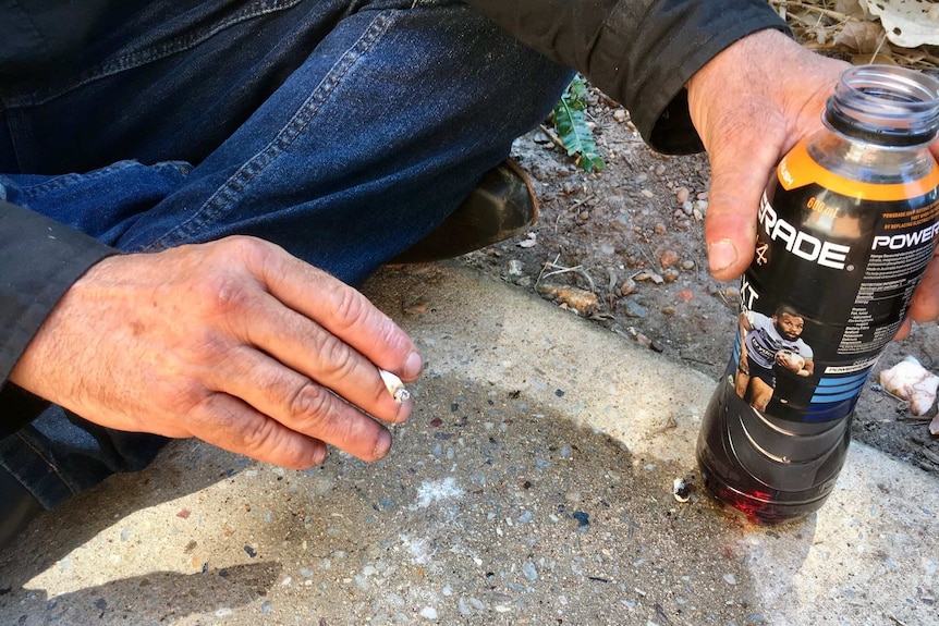A man's hands holding a cigarette and a plastic drink bottle filled with dark liquid