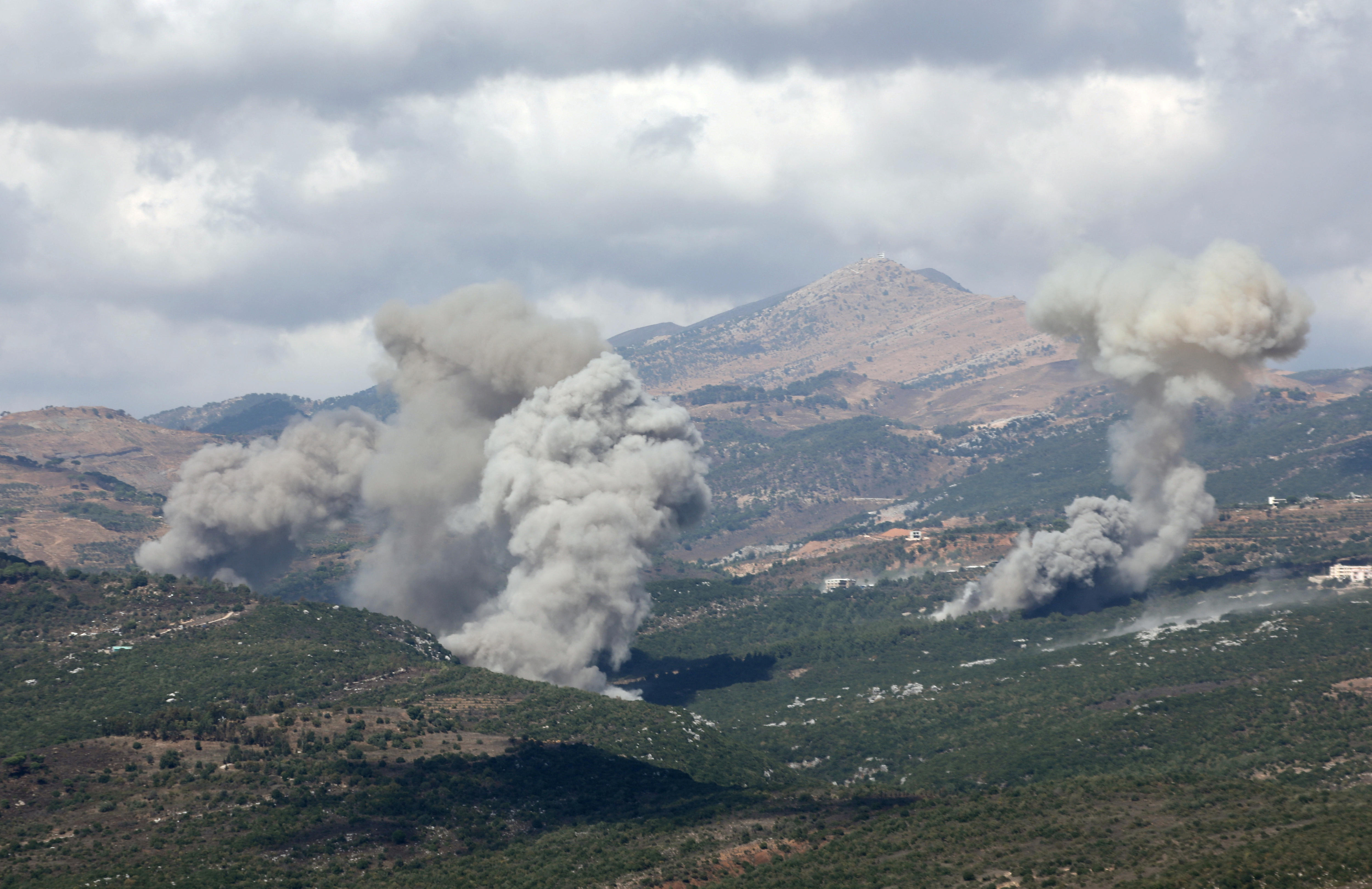 Smoke rises from tree-topped hillsides with homes dotted amongst the shrubbery against a cloudy sky