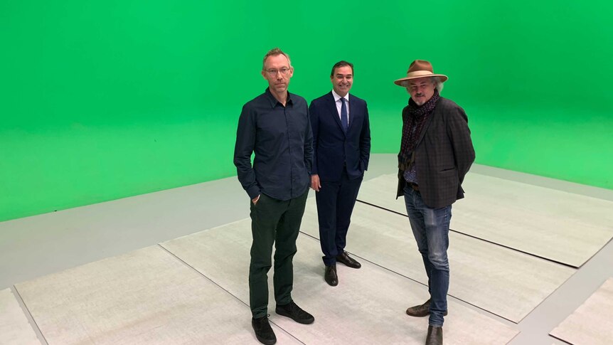 Three men stand in the middle of a room with a green screen behind them
