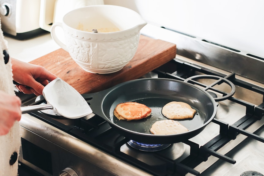 A person flips pikelets in a frying pan on a gas stovetop. The bowl of pikelet batter sits on the stove.