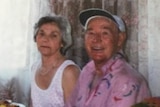 An older woman in a white singlet sitting next to an elderly man in a pink shirt and white cap.