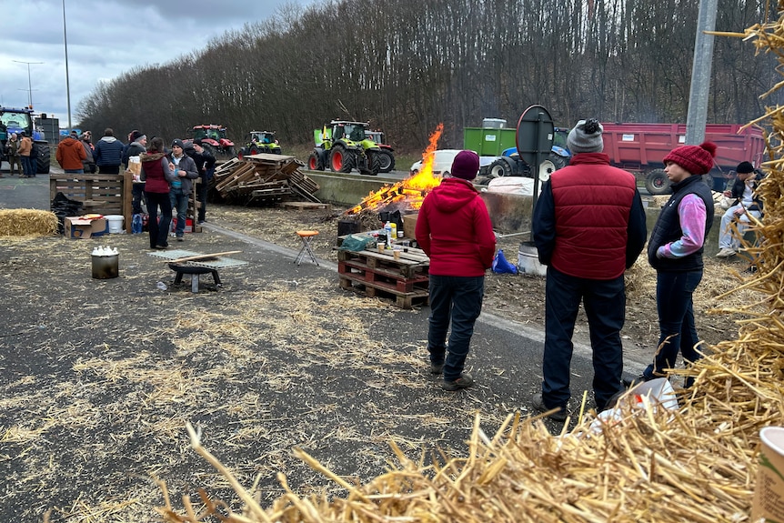 People stand on a highway surrounded by haybales, tractors and a small fire.