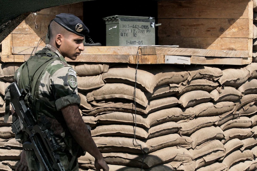 A French soldier dressed in camouflage and armed with a gun stands in front of stacked sandbags.