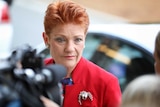 Pauline Hanson, wearing a red jacket with a silver brooch, looks past a camera in the foreground.