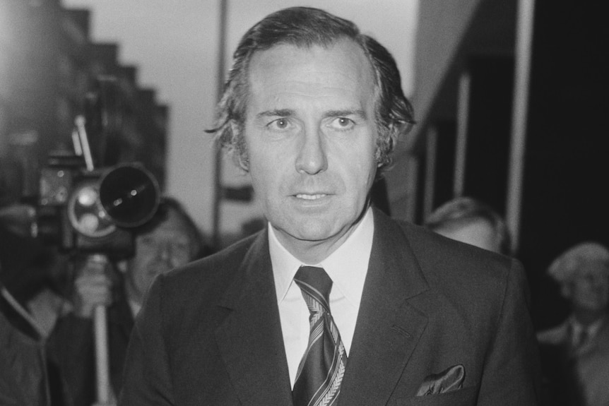 A black and white photo of John Stonehouse, a middle-aged man wearing a dark suit and tie, his mouth open slightly