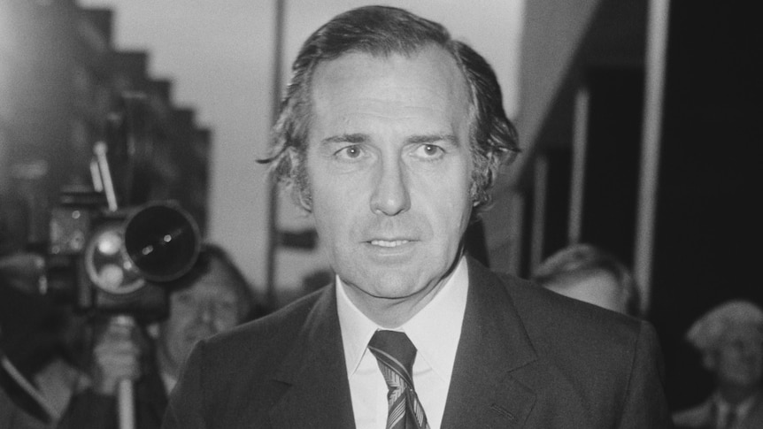 A black and white photo of John Stonehouse, a middle-aged man wearing a dark suit and tie, his mouth open slightly