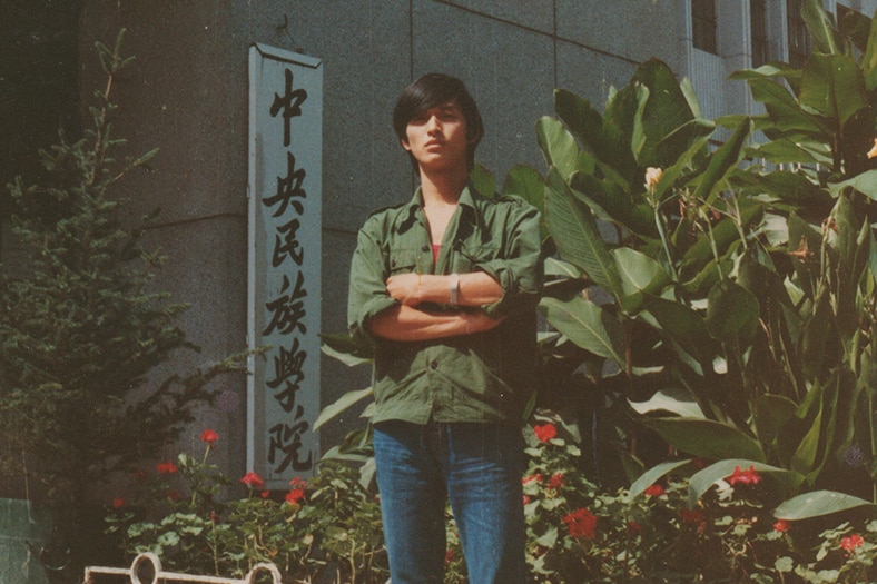 An old picture of a young Chinese man standing in front of a gate of a university.