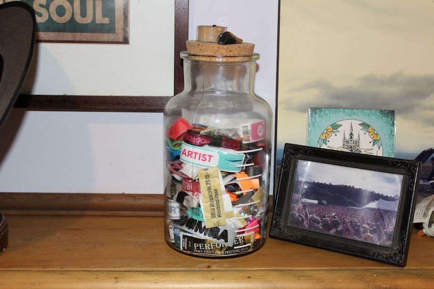 A large jar of festival wristbands sit on a wooden fireplace mantel alongside a picture of a crowd at a festival.