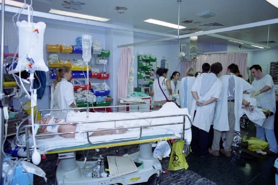 Man laying in hospital bed wrapped in blankets with doctors standing around 