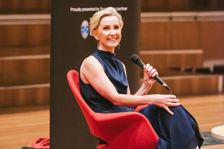 Deb Knight smiles as she sits in a chair holding a microphone on stage at a Women in Media function in Brisbane.