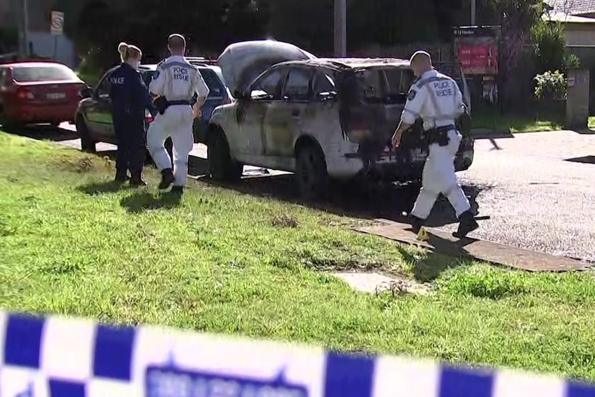 Police officers walking towards a burnt-out car.