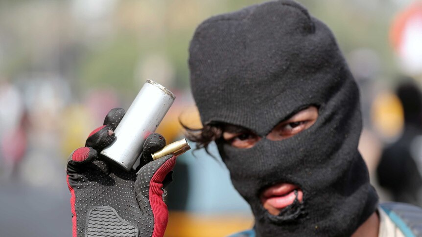 A man in a balaclava is seen wearing gloves and holding a tear gas and bullet canister.