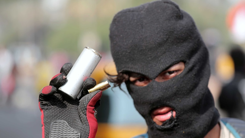 A man in a balaclava is seen wearing gloves and holding a tear gas and bullet canister.