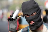 A man in a balaclava is seen wearing gloves and holding a tear gas and bullet canister.
