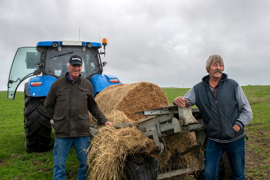 Two men stand next to a blue tractor carrying bales of hay in a green paddock with overcast skies.