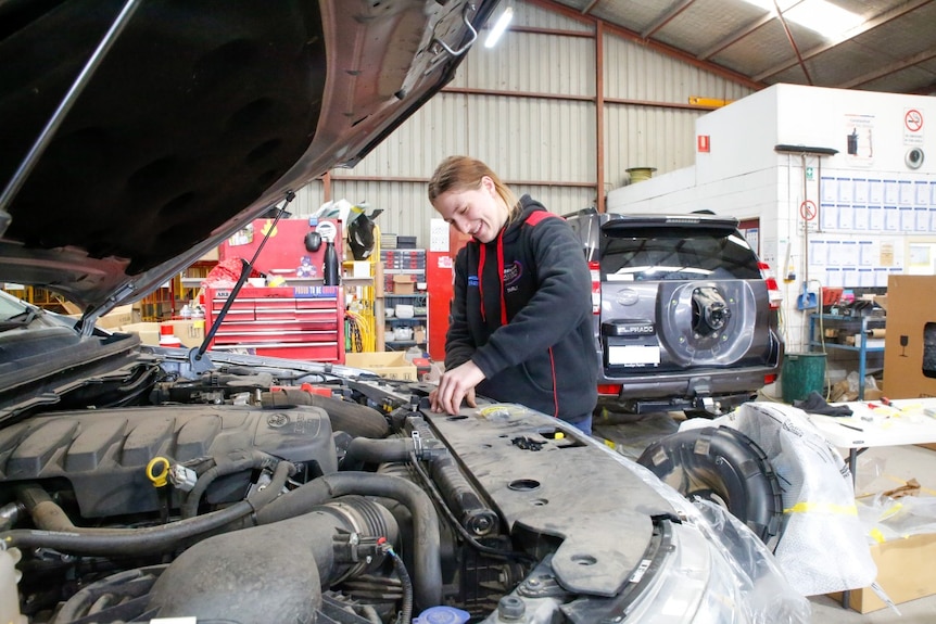 Woman works on a car with its bonnet open in a tin shed with another car in background.