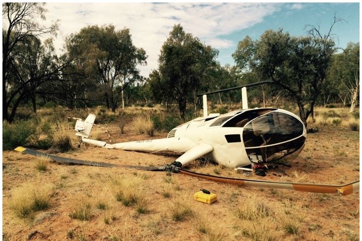 A close shot of a white helicopter on its side and on the ground. It is surrounded by arid bushland.