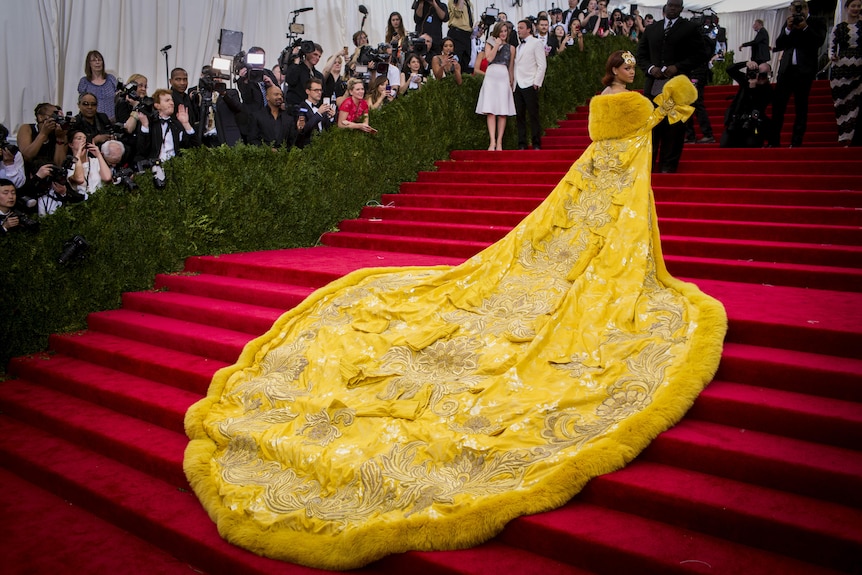 Rihanna wearing a yellow fur-lined gown with a long train stands on the red carpet at the Met Gala.