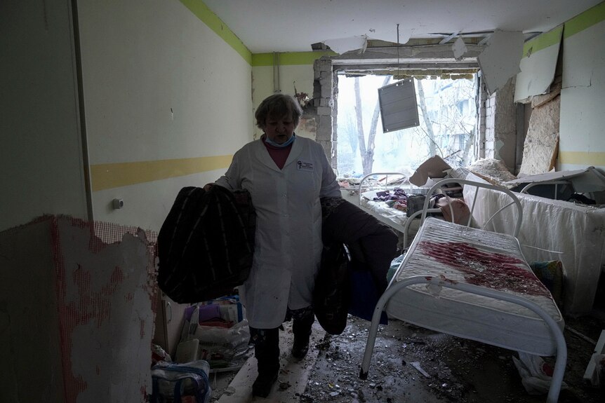 A medical worker inside a damaged hospital carries some supplies, near a bloodied single bed. A window and wall are blown out.