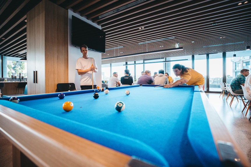 two men in T-shirts play pool on a pool table in an open office setting