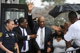 Bill Cosby stands among a group of people with his hand raised in the air to wave.
