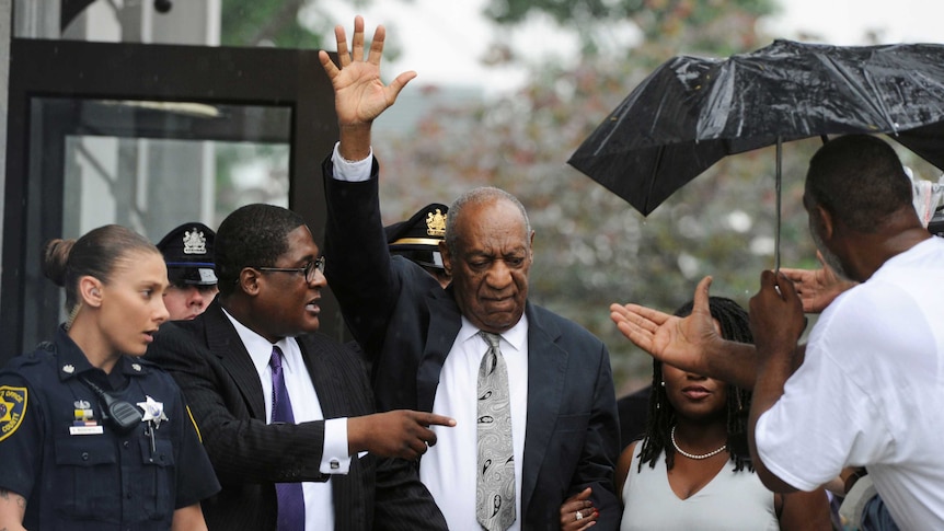 Bill Cosby stands among a group of people with his hand raised in the air to wave.