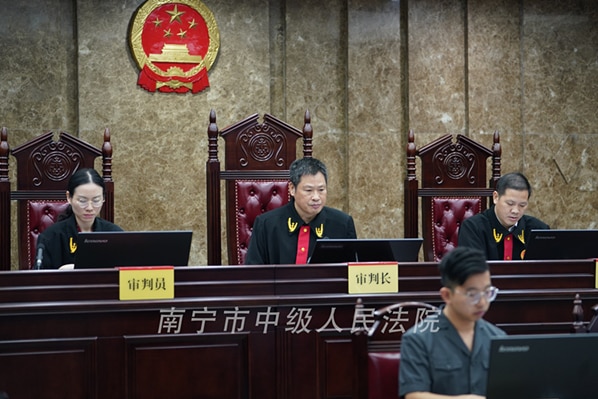 Three judges sit behind a tall mahogany desk on ornate chairs, while the Chinese national emblem is behind them on a brown wall.