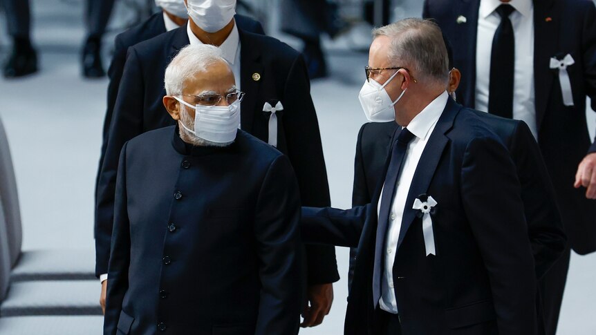 Two men in dark formal attire and face masks look at each other.