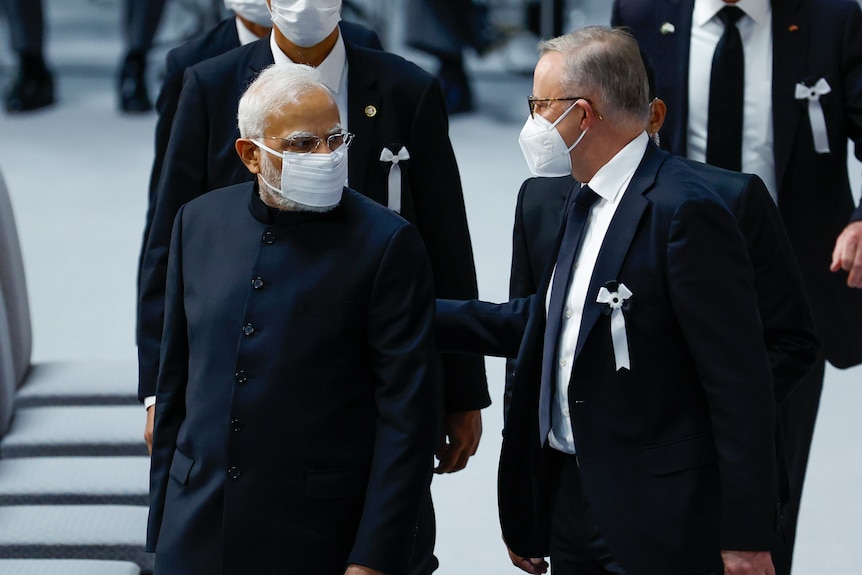 Two men in dark formal attire and face masks look at each other.