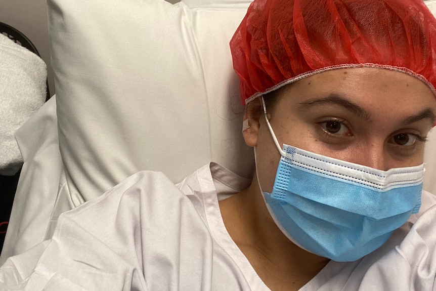 Ella Taverner wears a surgical mask, cap and gown, lying against a pillow in a hospital bed. 