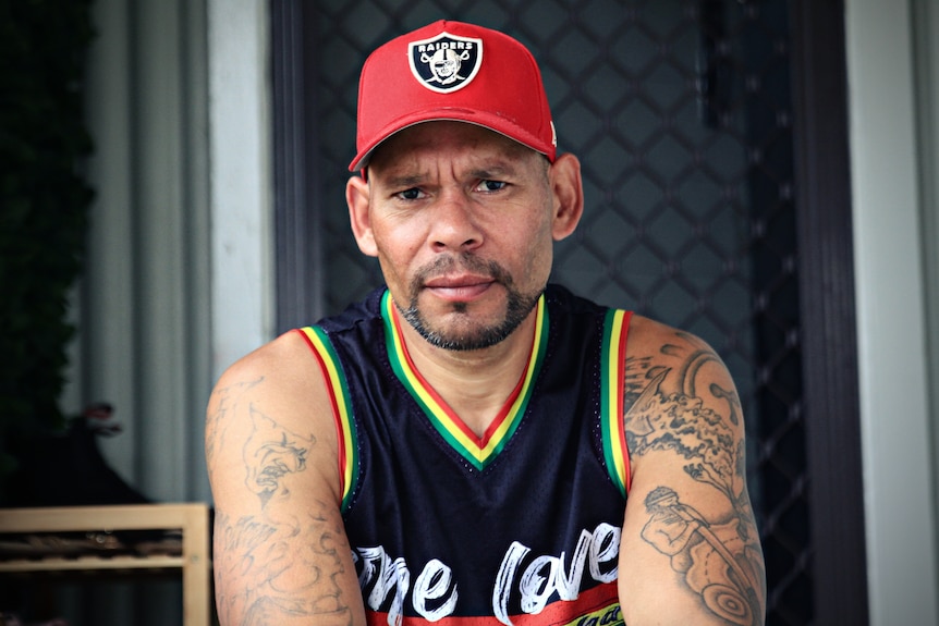 Man sitting down wearing a singlet and red cap. He has tattoos on his arms. 