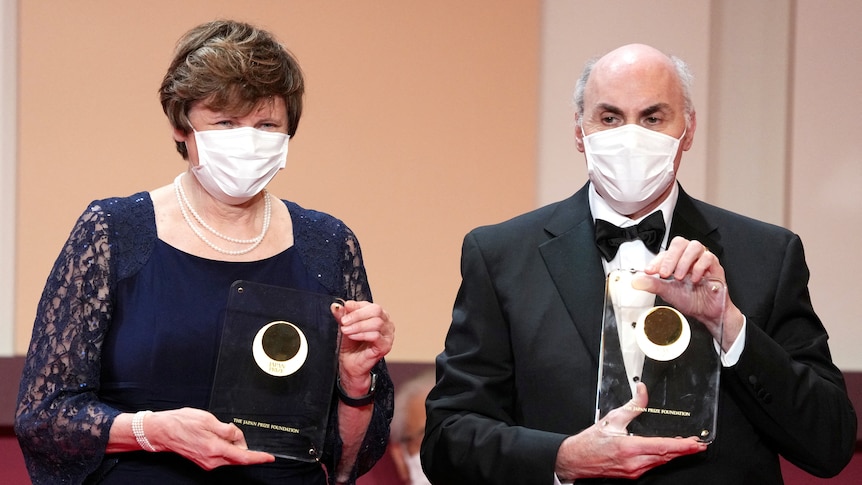 A man and a woman wearing masks and formal clothing pose with clear glass trophies.