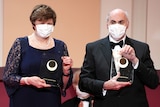 A man and a woman wearing masks and formal clothing pose with clear glass trophies.