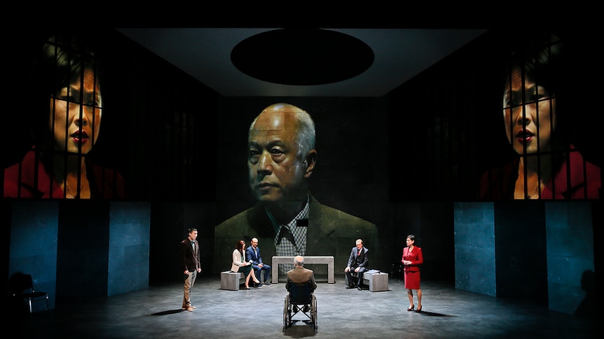 A man in wheelchair with back to audience faces two standing people, three seated on concrete benches and large projections.